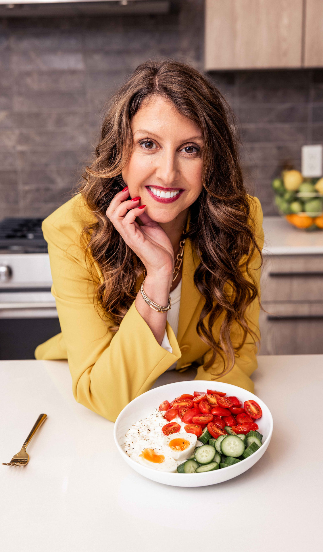 Simone Lovell in a bright yellow suit leaning over a bowl of cottage cheese, tomatoes, cucumbers, and eggs