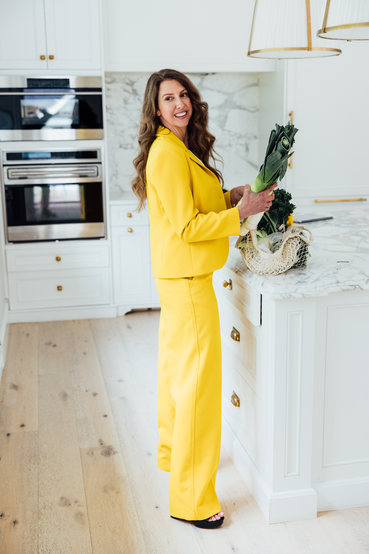 Simone Lovell in a bright yellow suit holding fresh greens in a sunlit kitchen